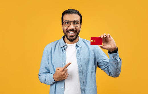 Happy man in a blue shirt proudly pointing at a red credit card in his hand, beaming with satisfaction, against a bright yellow background. Finance, money recommendation