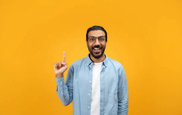 An excited man with a beard and glasses raises his index finger, sporting a bright smile, possibly sharing an idea or ready to speak, against a vivid orange background