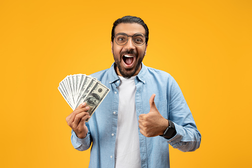 Ecstatic man in a blue denim shirt shows a fan of 100-dollar bills while giving a thumbs up, possibly celebrating financial success, winnings, or a good investment, on a yellow background