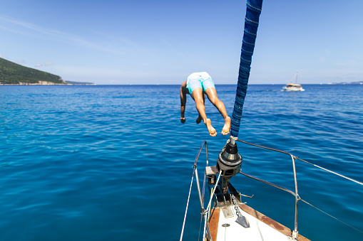 Man jumping into the sea from a sailboat. The sea is turquoise colored and crystal clear. He is wearing swimming trunks.