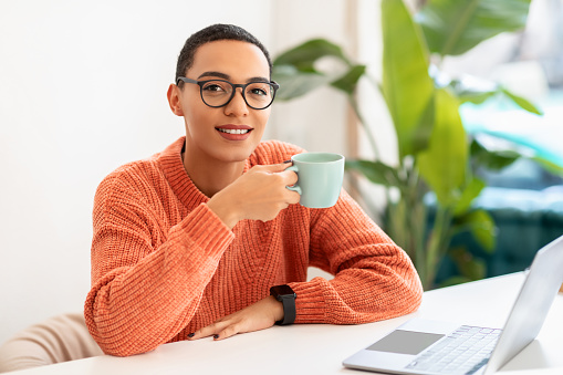 Happy calm young latin woman in glasses enjoy break and free time, drink cup of coffee at table with laptop in cafe, office interior. Create idea, relax, work and business
