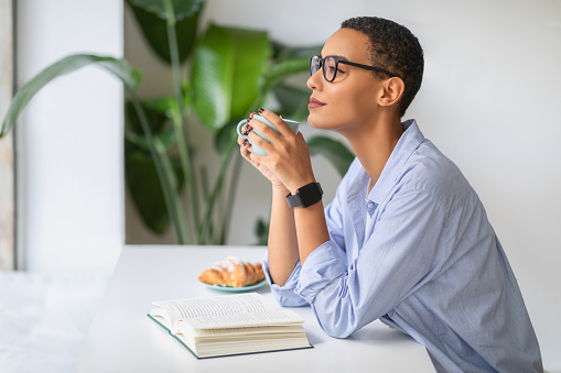 Relaxed happy latin young lady in glasses with notebook on table, enjoy book, cup of coffee in office, cafe. Morning routines, work breaks, peaceful reflection, study, planning day
