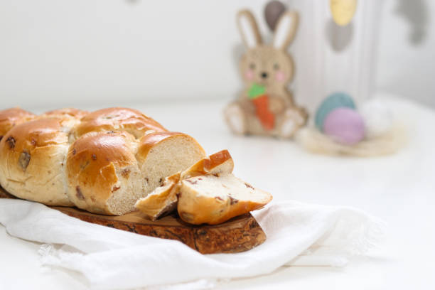 Cutted easter loaf bread on wooden plate on white table, blurry background with easter decoration.