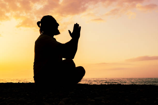 A man is sitting on the beach and praying. The sky is orange and the water is calm