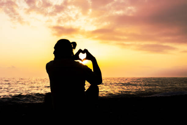 the man is sitting on the beach and making a heart shape with her hands. The sun is setting in the background, creating a warm and peaceful atmosphere
