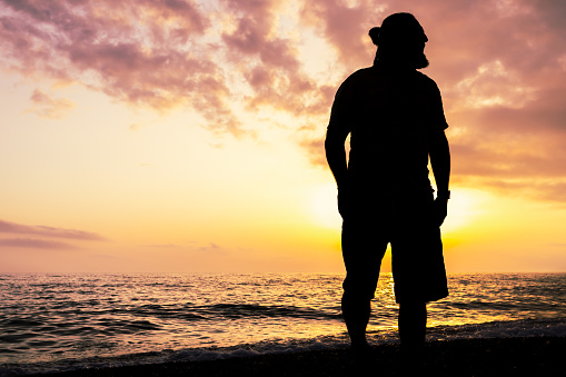 A man stands on a beach at sunset, looking out at the ocean. The sky is filled with clouds, creating a moody atmosphere. The man's silhouette against the backdrop of the ocean