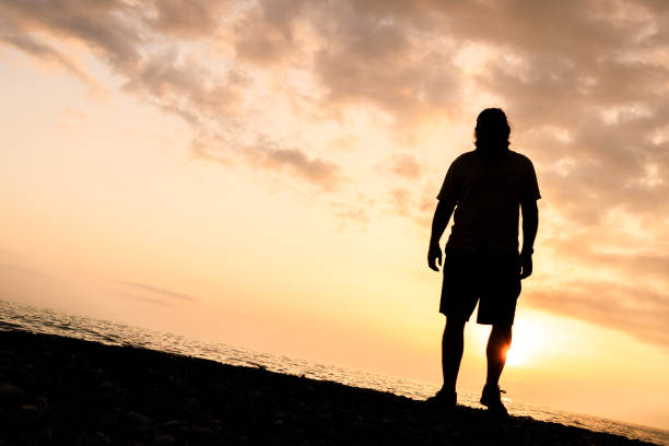istock A man stands on a beach at sunset, looking out at the water 2104683723