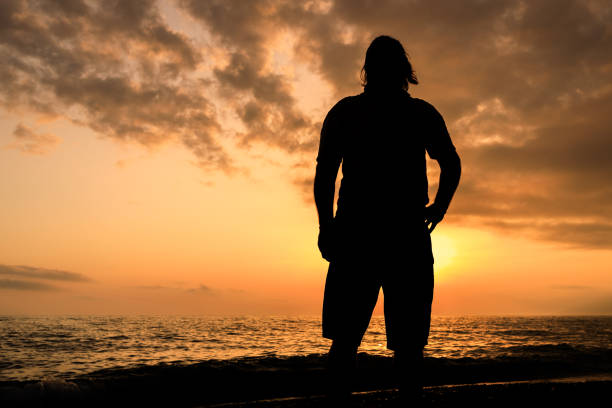 A man stands on a beach at sunset, looking out at the ocean. The sky is filled with clouds, creating a moody atmosphere. The man's silhouette against the backdrop of the sun