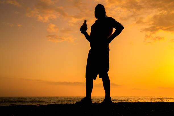 A man is standing on a beach at sunset, holding a bottle. Concept of relaxation and leisure, as the man enjoys his drink while watching the sunset over the water