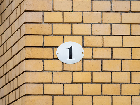 Number 1 on a yellow exterior house wall. The metal plate is enameled and white colored. The design very vintage and retro. The one is part of a residential address.