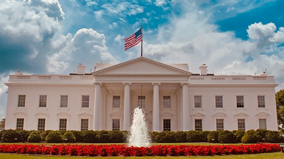 The South Portico of the White House. Washington DC. The White House is the official residence and principal workplace of the President of the United States, located at 1600 Pennsylvania Avenue. Beautifully landscaped lawn with flowers, fountain and blooming trees is in foreground. Deep blue clear sky is in background. American flag is flying atop. The image lit by spring evening sun. Canon 24-105mm f/4L lens.