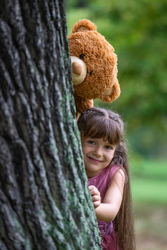 A sweet, pretty girl of 6 years old with long hair laughs cheerfully and looks out from behind a large tree. A large toy teddy bear peeks above her