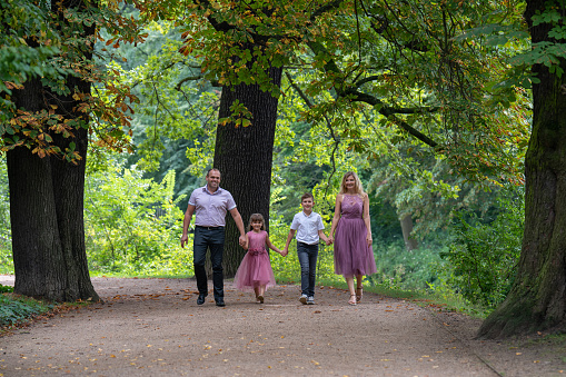 Portrait of a happy family. Laughing dad and mom, teenage son and first-grader daughter hold hands and walk along a path in the park. Mom and girl are dressed in beautiful pink dresses.