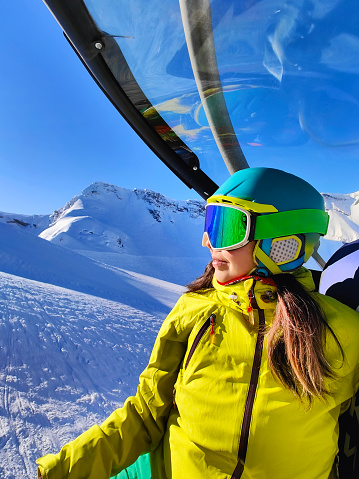 A skier gazes out from a ski gondola, the clear blue sky and snow-clad peaks reflecting in her goggles, capturing the anticipation of the slopes.