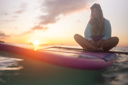 Portrait of a smiling woman sitting on a stand-up paddle board, floating on the sea and looking directly at the camera