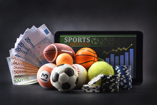 Concept of physical and online sports betting with balls representing various sports on a dark isolated background with banknotes, betting chips and tablet with betting application and sports analysis. Front view.
