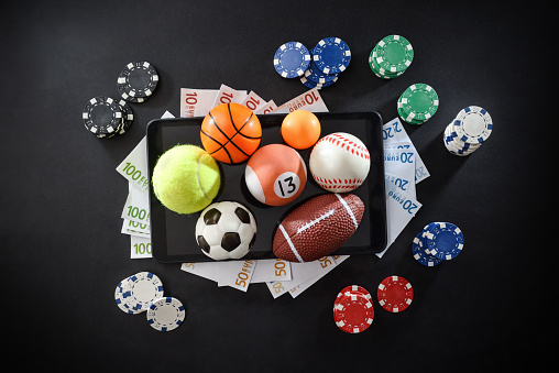 Online sports betting concept with various sports balls, money and chips on mobile devices isolated in the center on dark background. Top view.