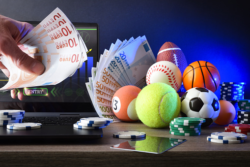 Person making online sports bets with laptop with application on desk with objects representing different sports, money and chips to bet. Front view.