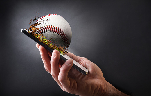 Concept of watching baseball online with hand holding a mobile phone with ball and dark gray isolated background.