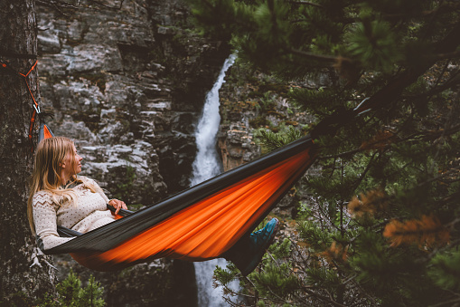 Summer vacations - woman chilling in hammock in forest travel outdoor tourist girl enjoying waterfall view in Norway adventure trip healthy lifestyle with camping gear weekend bivouac in wilderness