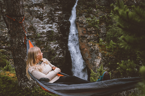 Woman enjoying waterfall view lying in hammock in forest travel lifestyle outdoor with camping gear summer vacations girl hiking in Norway eco tourism adventure trip in the wild