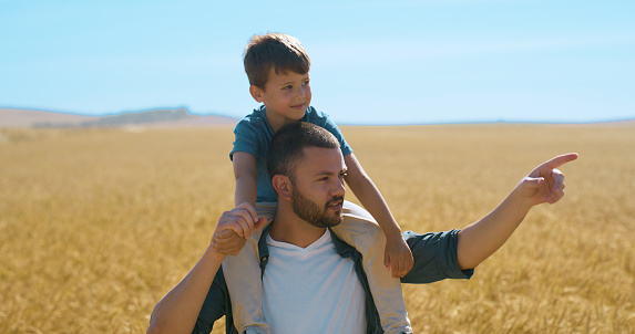 Wheat, farm and kid with piggyback on man for adventure, explore and vacation on field in Ukraine. Dad, son and sunshine with point for direction, location and teaching for childhood development