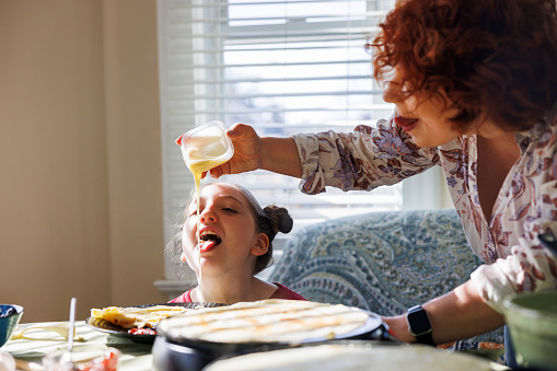 Little Caucasian girl with hair buns, tongue out, licking pancake filling during breakfast with mom. Redhead woman pouring condensed milk into the pancakes