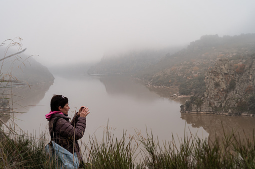 On a cold foggy morning, a woman stops on her hiking route to capture the beauty of the landscape with her smartphone. With the mighty Douro River meandering between rock-walled canyons as a backdrop, the atmosphere is mystical and enveloping. The limited visibility adds an aura of mystery to the scene, as the woman, focused on her task, seeks to capture the essence of this unique natural environment. Her silhouette is silhouetted against the misty landscape, highlighting her connection to nature and her passion for photography. Abelón, Castilla y León-Spain.