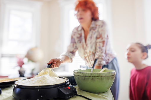 Blurred Caucasian redhead woman flips a pancake with a spatula on the electric skillet, her defocused daughter watching the cooking process in the sunny kitchen. Focus on a pancake on electric pan