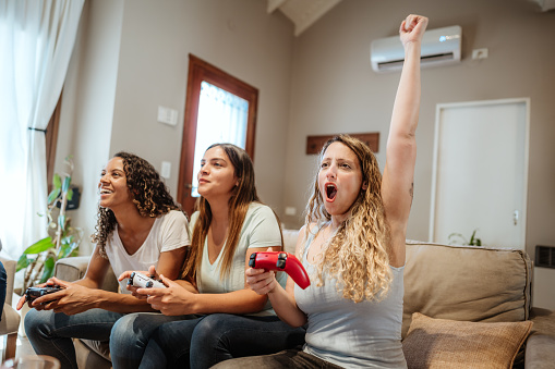 Three women sit on a sofa, joyfully playing video games with one celebrating a win. The excitement and competition are palpable.