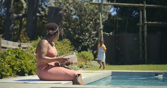 Swimming pool, summer and black woman with kid in backyard for development, bonding and afternoon enjoyment. African people, poolside and relax with book for story, literature novel or poetry