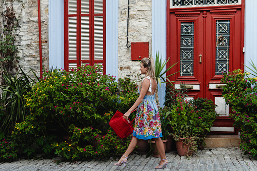 Young Woman Strolling Past Historic House with Red Doors and Bunch of Different Plants and Flowers in Mediterranean Village.