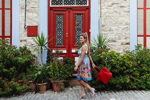 Young Woman Strolling Past Historic House with Red Doors and Bunch of Different Plants and Flowers in Mediterranean Village.