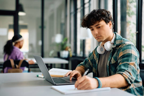 Young student taking notes while e-learning on laptop at the university. University student writing while using laptop and studying in the classroom. Copy space. education stage stock pictures, royalty-free photos & images