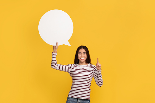 Cheerful good looking asian lady smiles gladfully points up, shows white communication bubble, mockup and space for social media announcement or news, share idea, isolated over yellow wall