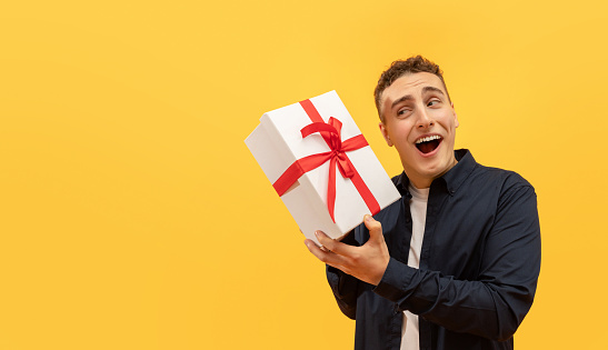 Excited young guy holding white gif box with red bow next to his ear, wondering what is inside, isolated on yellow background. Birthday man enjoying his presents, panorama with copy space