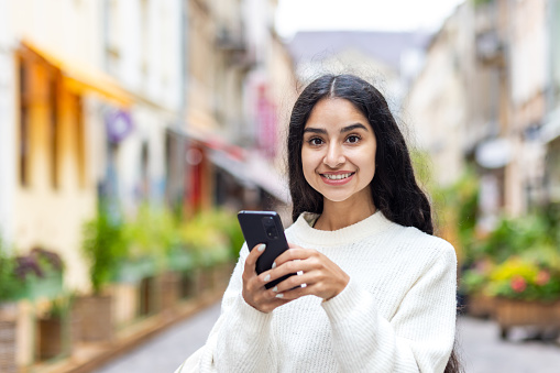 Retrato de una niña india sonriente y hermosa de pie en medio de una calle de la ciudad, sosteniendo un teléfono móvil y mirando a la cámara photo