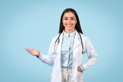 Smiling female doctor presenting something invisible with one hand, wearing stethoscope and white lab coat, standing confidently against calming blue background