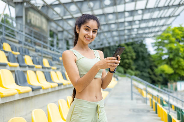 mujer india activa con reloj inteligente usando teléfono inteligente en el estadio - estilo de vida fitness -   brunette  fotografías e imágenes de stock