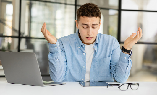 Unhappy tired millennial european guy manager spreads hands, suffer from mistake, problems at table in coworking office. Stress at work, business, deadline, bad job, burnout