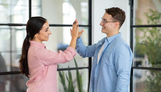 Glad millennial arab lady and european guy manager giving high five with hands, celebration victory, success in coworking office, panorama. Motivating, teamwork, business together, lifestyle