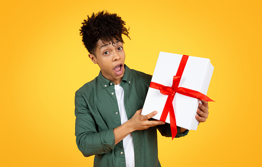 Amazed handsome stylish young african american guy holding white gift box with red ribbon and grimacing, got birthday present, isolated on yellow studio background