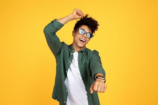 Carefree stylish young black guy wearing sunglasses with smart watch on his wrist dancing and singing isolated on yellow studio background, have fun, enjoying holiday or vacation