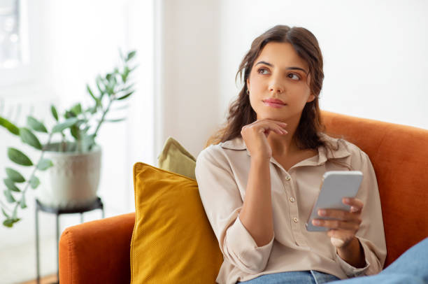 Woman holding smartphone and looking away while relaxing on couch at home Pensive young woman holding smartphone and looking away while relaxing on couch at home, beautiful female deep in thoughts using mobile phone in domestic interior, thinking and planning something contemplation stock pictures, royalty-free photos & images