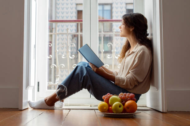 istock Serene young woman reading book beside bright window at home 2105212037
