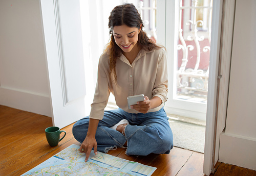 Young woman using her smartphone and city map to plan journey, sitting on wooden floor by the window at home, happy millennial female showcasing adventure preparation, getting ready for trip
