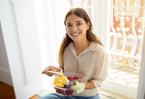 Happy young woman holding bowl of colorful mixed fruits and smiling at camera, beautiful european female enjoying healthy snack while sitting on floor by the window at home, closeup shot
