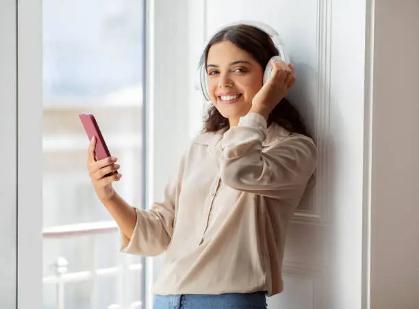 Happy young woman wearing wireless headphones enjoying listening music on smartphone, smiling beautiful female standing by the window in sunny room, enjoying relaxing at home, free space