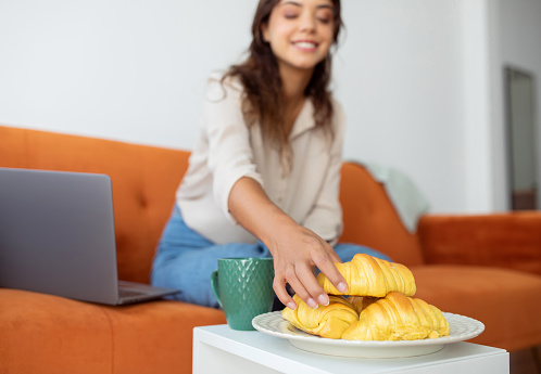 Smiling woman reaching for croissants on plate next to her laptop, happy young female taking tasty pastry, having break while working with computer in living room at home, selective focus
