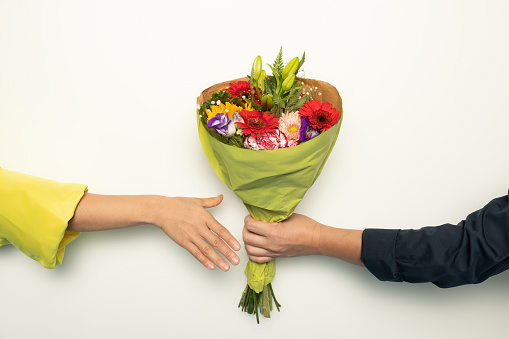 Two hands, one clad in a yellow sleeve and the other in a dark blue sleeve, are about to touch, sharing a vibrant bouquet of flowers wrapped in green paper, cropped, close up